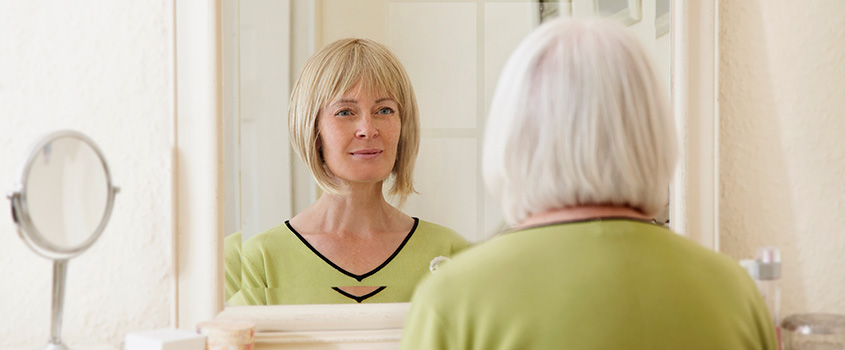 Ältere Frau schaut in den Spiegel. Verlinkung zum Anfang der Bestellung.