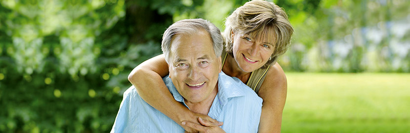 Seniorin ummarm ihren Mann von hinten und beide lächeln glücklich in die Kamera